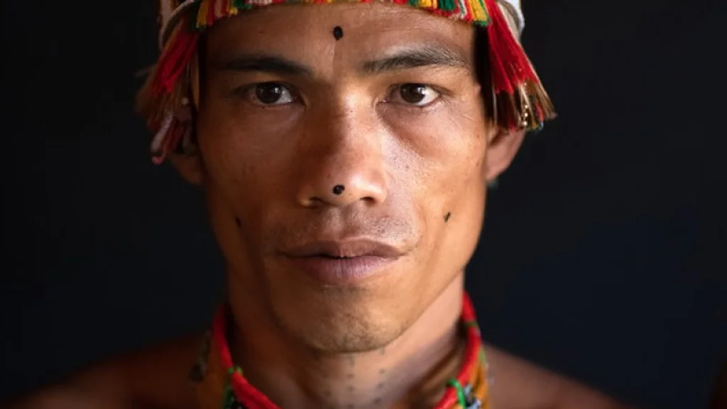 A close up portrait of an Indigenous Mentawai man with traditional facial markings and a colorful woven headband against a dark background. He gazes directly at the camera with a serious expression, highlighting the cultural identity and resilience of forest-dwelling communities.