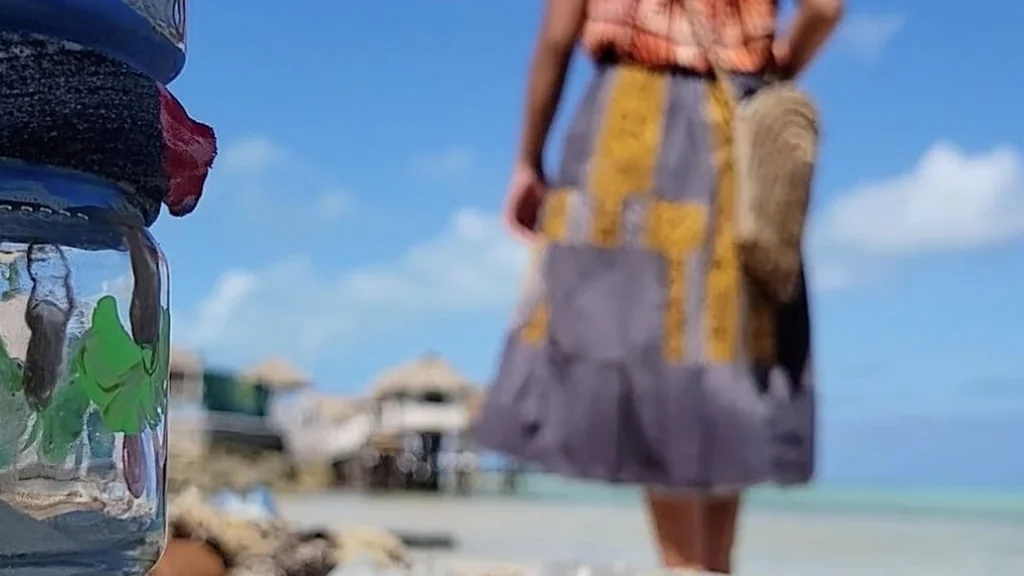 A low-angle shot features a glass jar with green painted details in the foreground. In the blurry background, a person in a patterned skirt and orange top stands on a sunny beach under a clear blue sky, near coastal structures and turquoise water.