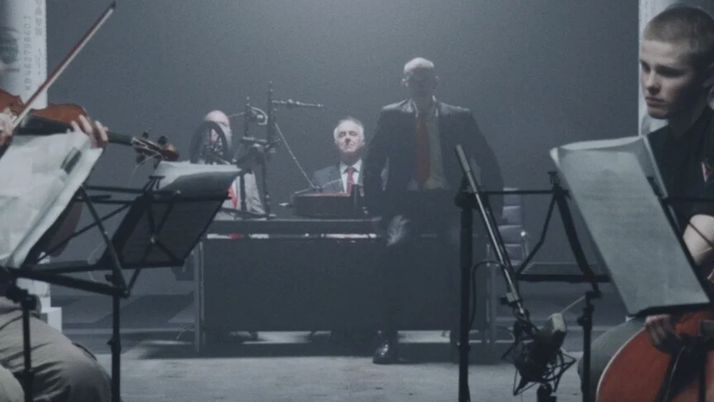 A dimly lit still shows a concert hall. Musicians with violins and cellos are playing in the foreground. Two men in suits are standing in the back, one behind a desk and another one standing up.