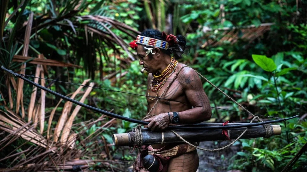 An Indigenous Mentawai man with traditional tattoos and headgear stands in a lush green jungle. He holds a large black bow and carries a bamboo quiver across his back, embodying a traditional hunter-gatherer lifestyle amidst dense tropical foliage and palm fronds.