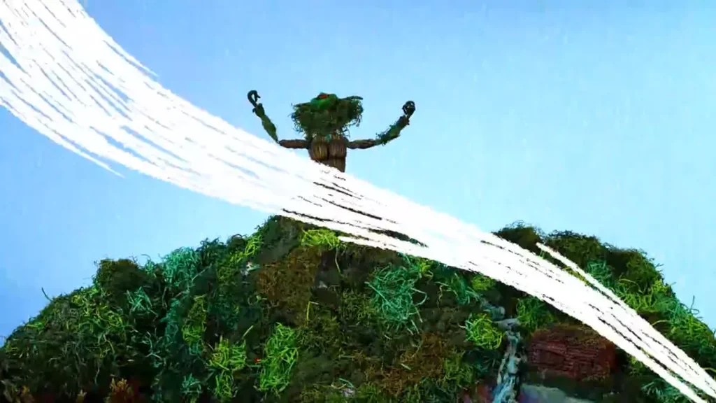 Μια φιγούρα stop-motion φτιαγμένη από φυσικά βρύα και ξύλο στέκεται στην κορυφή ενός πράσινου βουνού με τα χέρια υψωμένα. Πυκνές λευκές πινελιές διαγράφουν τόξα στο κάδρο πάνω από τον καθαρό μπλε ουρανό, υποδηλώνοντας άνεμο ή πνευματική ενέργεια που εκπορεύεται από τον γήινο χαρακτήρα.