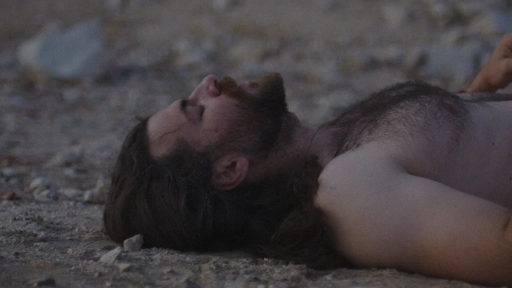 A close up profile shot of a man with a dark beard and long hair lying flat on his back on dry, rocky ground. The lighting is dim and somber, emphasizing the harsh, arid texture of the earth beneath him in a desolate landscape.