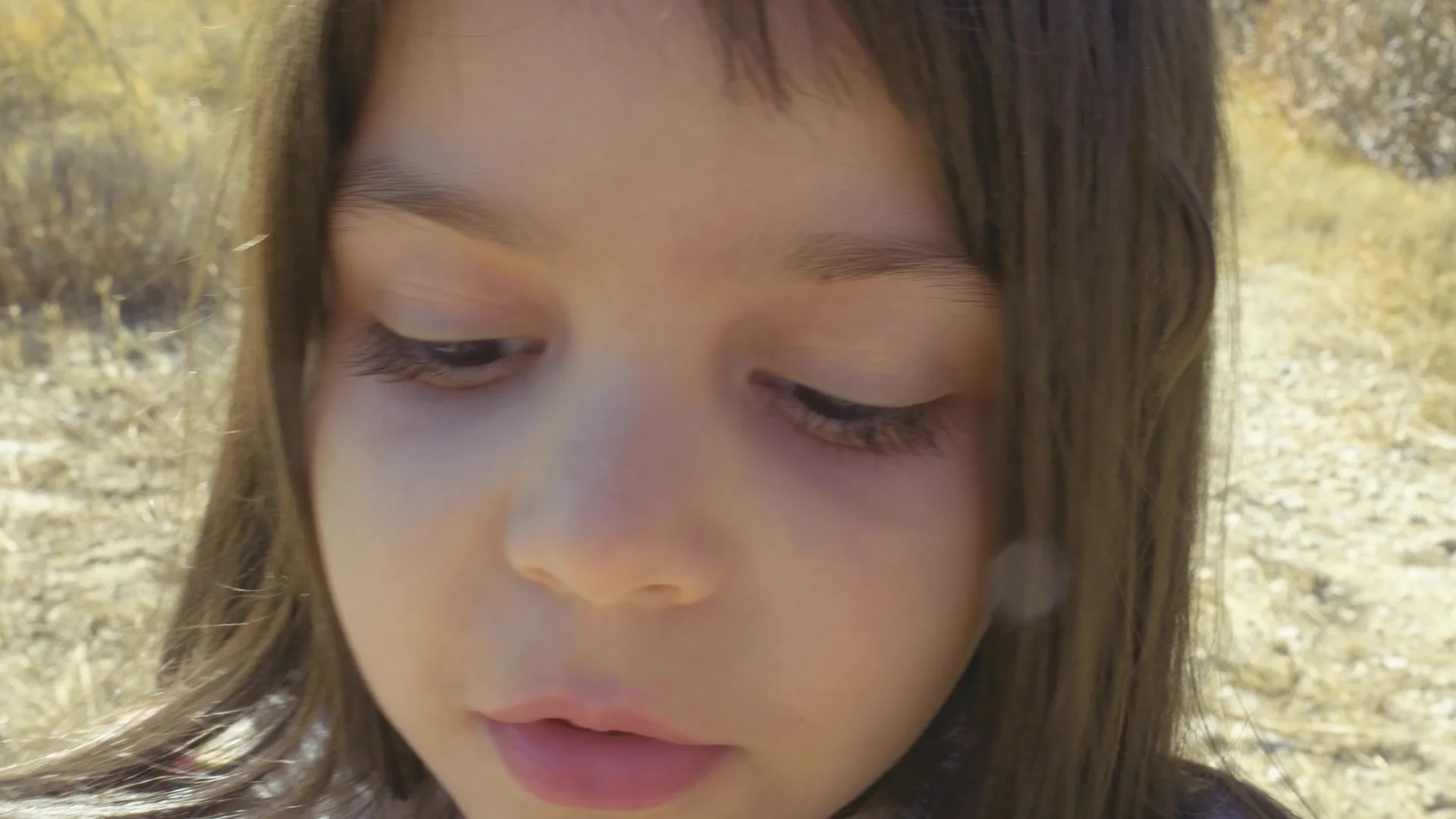 A close-up shot of a young girl with brown hair looking downward with a somber expression. She is outdoors in a dry, sunlit environment with blurred vegetation in the background, conveying a sense of vulnerability and reflection in a harsh landscape.