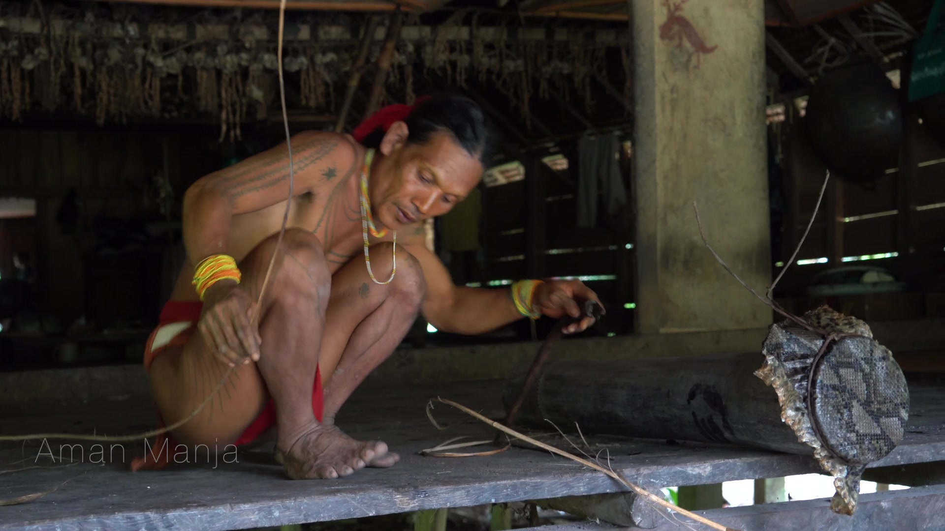 Still from "Mentawai - Souls of the Forest": An aerial view shows a traditional longhouse with a thatched roof in a clearing by a river, surrounded by dense green rainforest. The film title is overlaid.