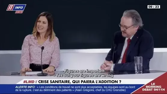 Στιγμιότυπο από το "The Flood": Μια γυναίκα με ροζ πουκάμισο χειρονομεί ενώ μιλά σε έναν άνδρα με κοστούμι και γυαλιά, και οι δύο καθισμένοι σε ένα σκούρο τραπέζι κατά τη διάρκεια μιας συζήτησης. Αγγλικοί υπότιτλοι είναι ορατοί.