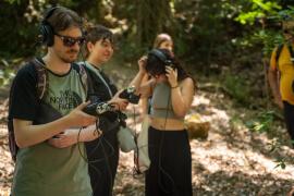 Participants in a sound recording workshop in a forest, using professional field recorders and wearing headphones to capture ambient nature sounds.