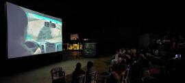 An outdoor film screening at night in a village, with an audience watching a movie on a large screen and the Solar Cinema van in the background.